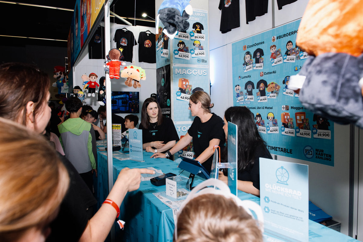 Kassenbereich des LunyMerch Stands auf der gamescom 2026 mit Team, Preisplakaten der Creator und Glücksrad Aufsteller