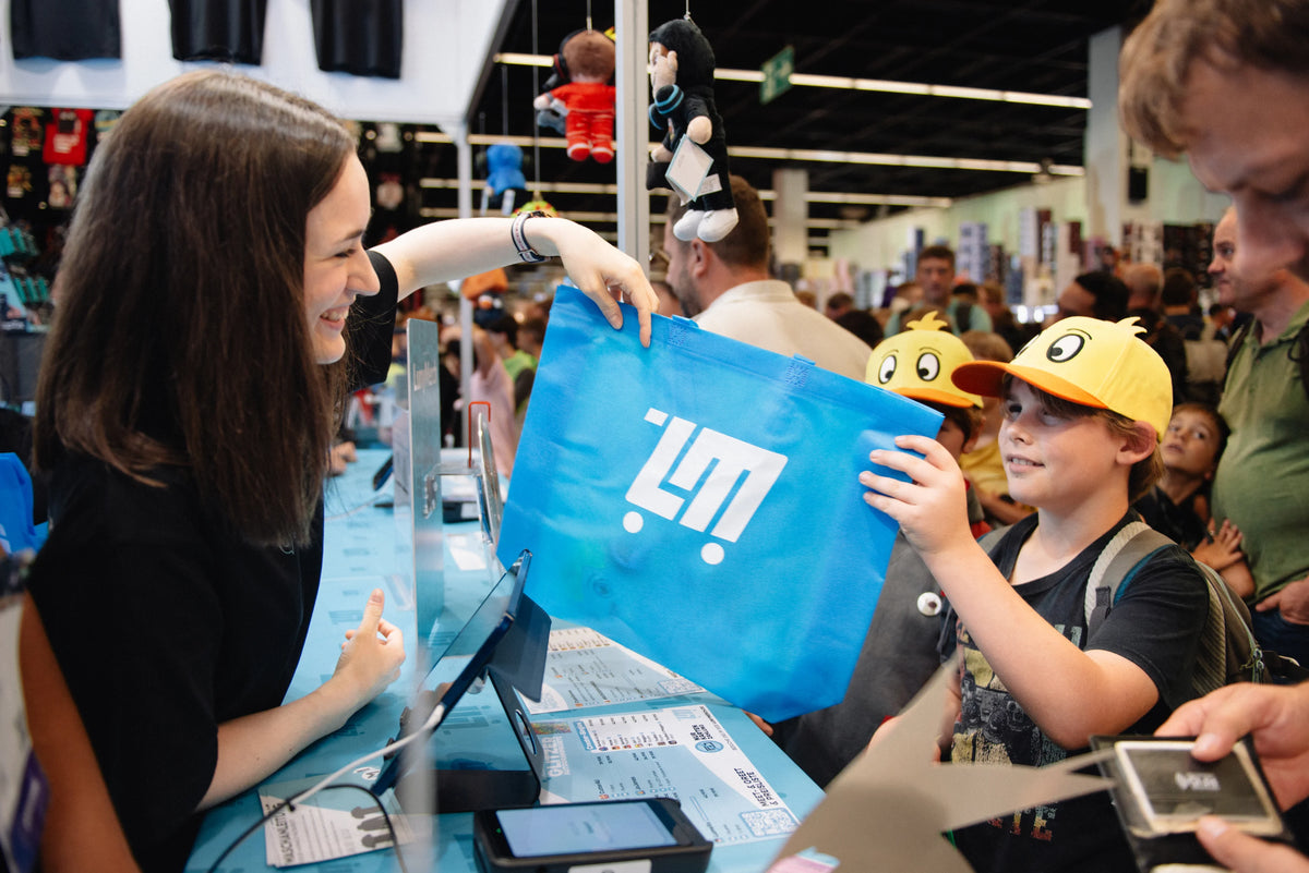 Mitarbeiterin überreicht einem jungen Fan eine blaue LunyMerch Tragetasche am Messestand auf der gamescom 2026